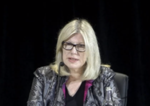A photo of a middle aged woman with light skin and blond mid-shoulder length straight hair. She is wearing dark framed glasses and a silver jacket over a black and pink top. She is speaking and looking to the her left side.